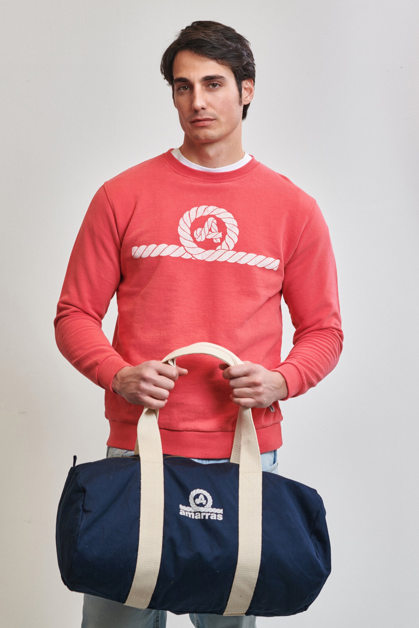 Bolsa tipo duffle de lona azul marino con asas de algodón crudo y logotipo clásico de Amarras, llevada por un modelo con sudadera roja.