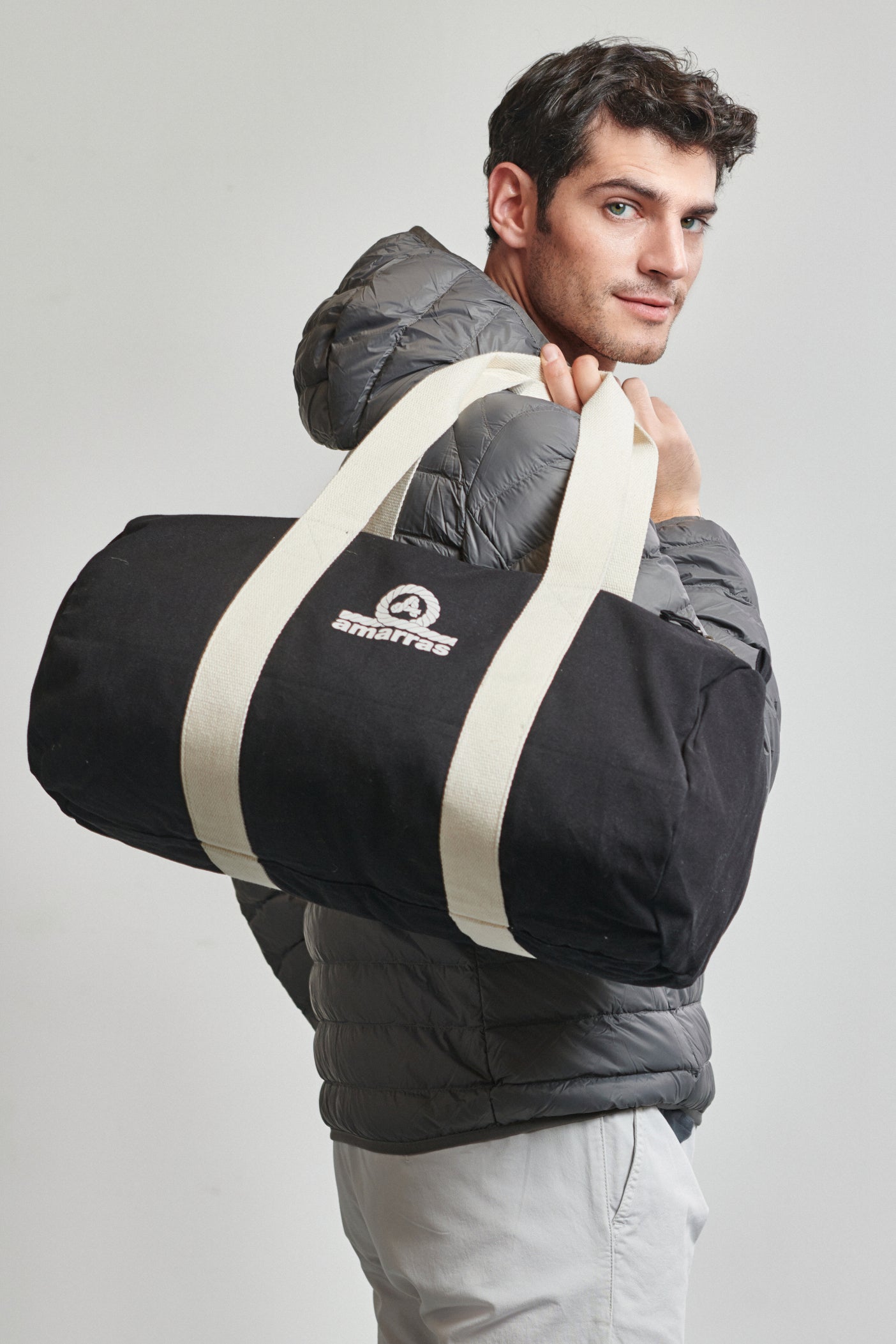 Bolsa tipo duffle de lona azul marino con asas de algodón crudo y logotipo clásico de Amarras, llevada por un modelo con chaqueta ligera de plumas Staff gris.