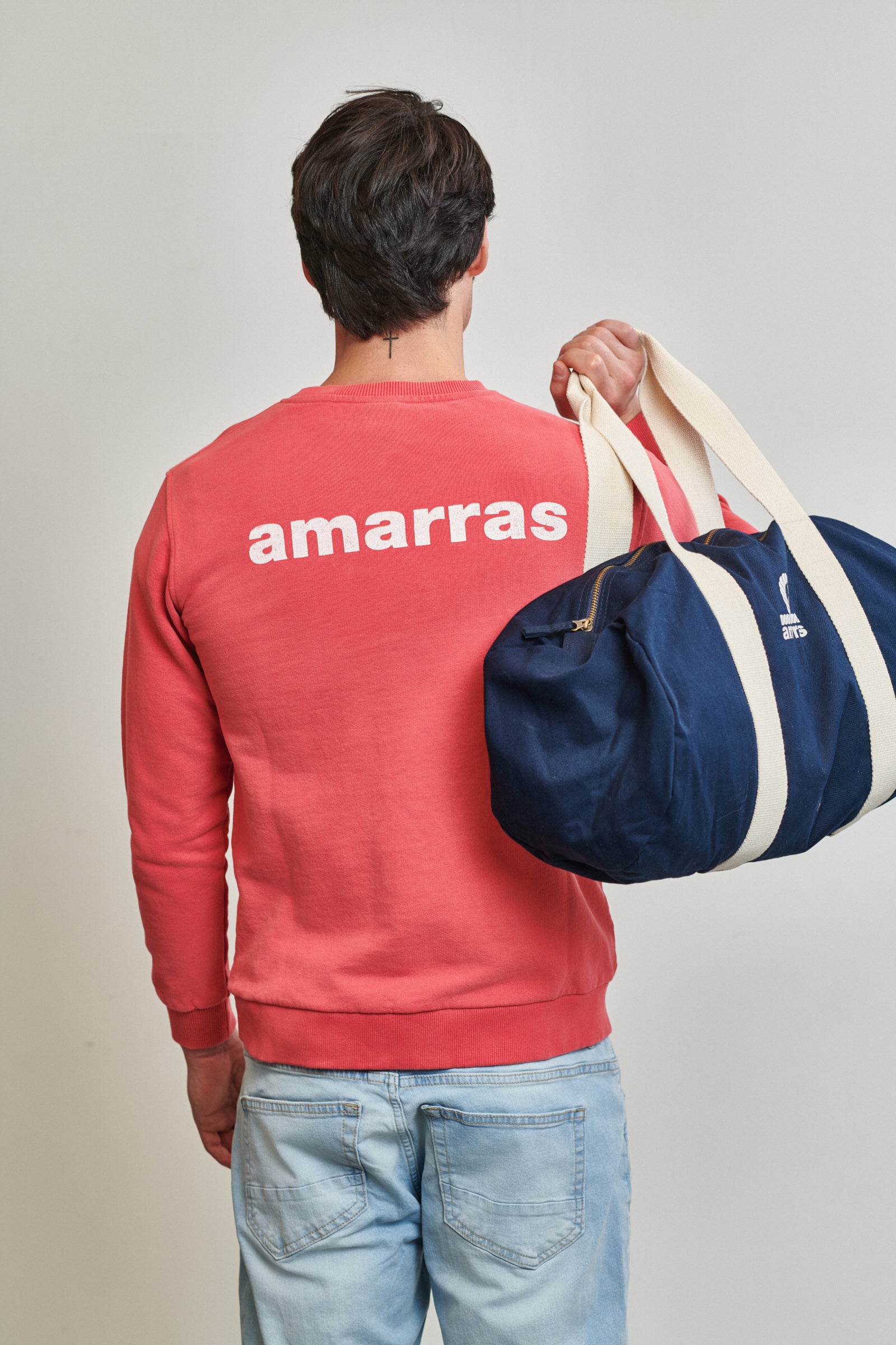 Bolsa tipo duffle de lona azul marino con asas de algodón crudo y logotipo clásico de Amarras, llevada por un modelo con sudadera roja.